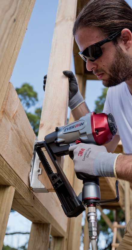 Close-up of a worker operating a joist hanger nailer to secure a metal hanger at a wood joint, demonstrating the tool's use in tight spaces.
