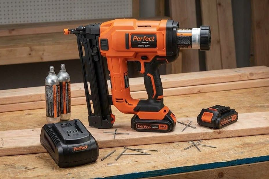 A gas powered nail gun set on a wooden workbench next to a battery charger, spare batteries, and fuel cells.